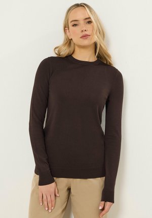 Donna bionda che indossa un maglione marrone scuro a maniche lunghe e pantaloni beige, in piedi contro una parete chiara.