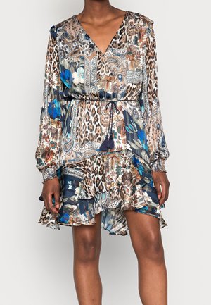 Femme portant une robe à manches longues, col en V, avec un mélange d'imprimés animaux et des motifs floraux bleus, taille cintrée et jupe asymétrique à volants.