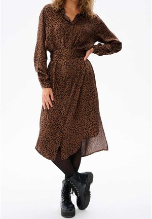 Frau trägt braunes knöpfbares Midikleid mit schwarzem abstraktem Muster, schwarze Strumpfhosen und schwarze Plateaustiefel mit Schnürung und posiert mit einer Hand in der Hüfte.