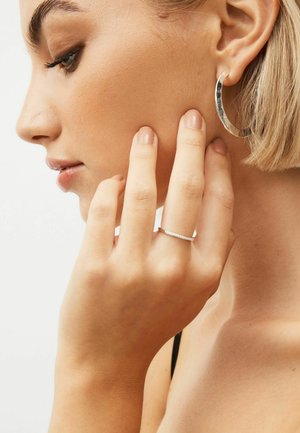 Silver hoop earring and a thin, sparkling ring on a hand, showcasing smooth skin and neutral nail polish against a light background.