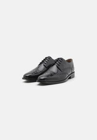 Un paio di eleganti scarpe da uomo in pelle nera lucida con dettagli brogue e alla francese su sfondo bianco.