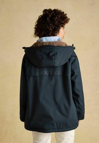 Tmavý petrolejový kabát s kapucňou, s hnedým fleecovým podšitím a viditeľným logom na chrbte. Tkanina pôsobí hladko a je odolná proti vode.