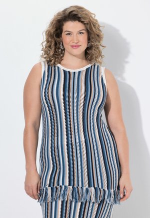 Frau mit lockigem Haar, die ein ärmelloses, blau-weiß-beige gestreiftes Strickoberteil mit Fransen am Saum trägt und vor einem weißen Hintergrund steht.