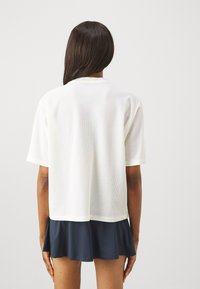 Top alb din plasă cu mânecă scurtă, cu decolteu rotund și o croială lejeră, purtat peste o fustă de culoare bleumarin închis. Textură netedă cu un design respirabil.