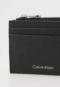 Black textured card holder with a zip closure, featuring multiple card slots, and a silver "Calvin Klein" logo on the front.