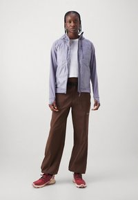 Gaiši violeta rāvējslēdzēja jaka ar divām priekšējām kabatām, valkāta pāri baltam topam. Brūnas vaļējas bikses un sarkani sporta apavi. Gludas faktūras.