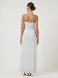 Γυναίκα με τα μαλλιά σε κότσο φοράει ένα μακρύ άσπρο φορεμα με παγιέτες και λεπτές τιράντες, στέκεται με την πλάτη προς το κοινό, σε ένα απλό φωτεινό φόντο.