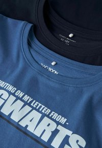 Dos camisetas de algodón, una de color azul marino y otra azul claro, exhibidas con cuellos redondos. La camiseta azul presenta un texto blanco: "ESPERANDO MI CARTA DE-".