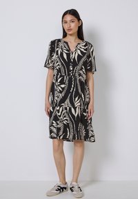 Schwarzes Kurzarmkleid mit weißem botanischem Muster, V-Ausschnitt und gerafftem Rock. Kombiniert mit hellen Sneakers, vor einem schlichten Hintergrund.