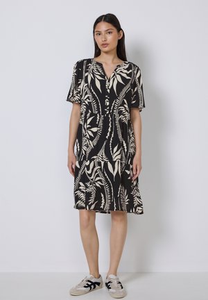 Schwarzes Kurzarmkleid mit weißem botanischem Muster, V-Ausschnitt und gerafftem Rock. Kombiniert mit hellen Sneakers, vor einem schlichten Hintergrund.