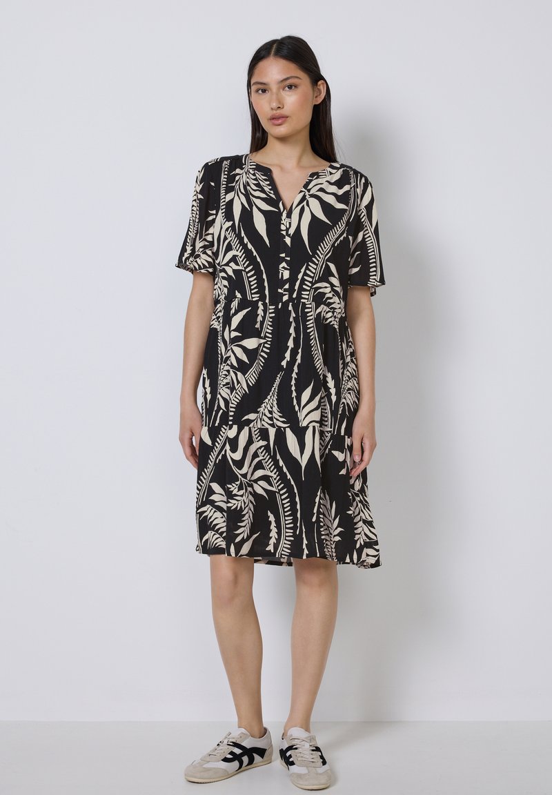Schwarzes Kurzarmkleid mit weißem botanischem Muster, V-Ausschnitt und gerafftem Rock. Kombiniert mit hellen Sneakers, vor einem schlichten Hintergrund.