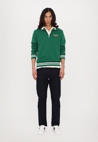 Grønn genser med glidelås på kragen, hvite striper i nederkant og på mansjettene, kombinert med mørke jeans og hvite sneakers. Casual sportdesign.