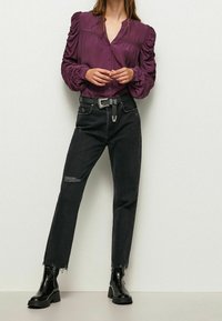 Camicia a maniche lunghe con bottoni di colore viola scuro, con maniche a sbuffo; abbinata a jeans neri strappati e stivaletti neri.