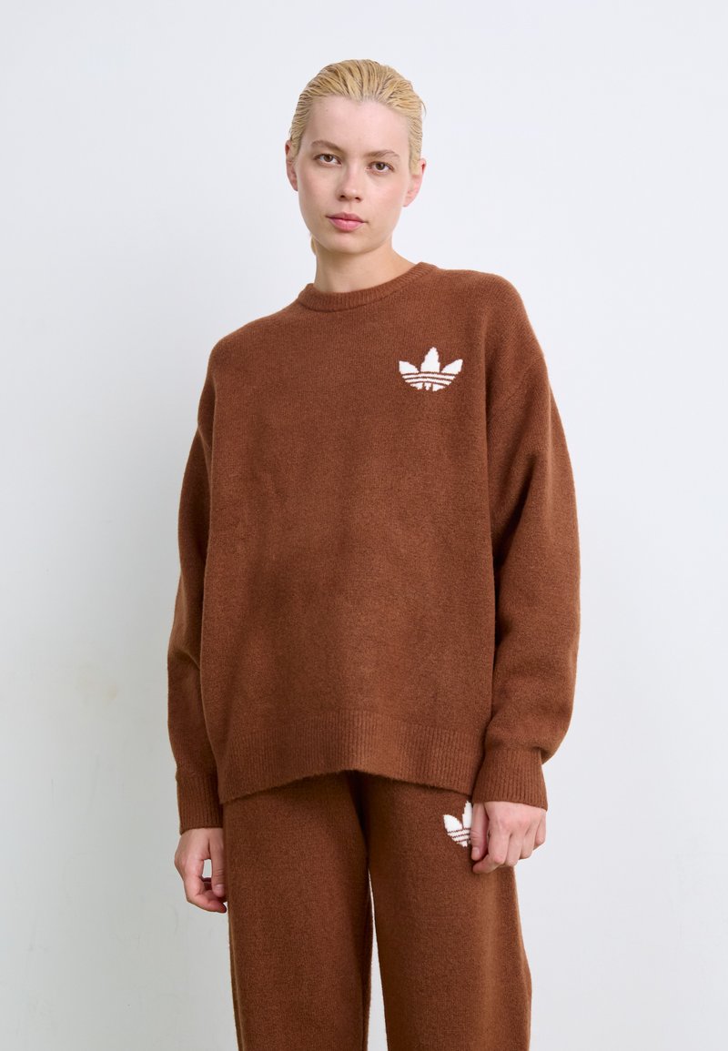 Νεαρό άτομο με ξανθά μαλλιά που φοράει καφέ πλεκτό πουλόβερ και παντελόνι με λευκά λογότυπα Adidas, στέκεται μπροστά σε απλό λευκό φόντο.