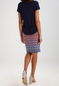 Granatowa bluzka z krótkim rękawem w połączeniu z spódnicą do kolan w czerwono-biało-niebieski wzór w zygzak. Jasnoszare buty na wysokim obcasie.