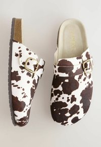 Zuecos de estampado de vaca en marrón y blanco con un hebilla en tono dorado y una plantilla acolchada. Parte superior texturizada y suela negra sólida.