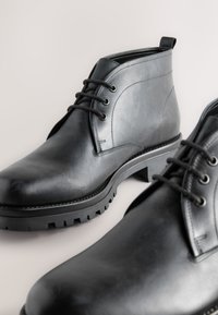 Schwarze Leder-Stiefeletten mit Schnürsenkeln, Gummisohlen mit Profil, glatter Oberfläche, abgerundeter Spitze und kleinem Zuglasche hinten für leichtes Anziehen.
