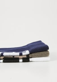 Quatre paires de chaussettes empilées horizontalement : marine, beige, blanche et noire. Texture tricotée avec des rayures de couleur contrastée en haut.