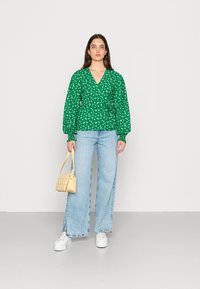 Blusa verde floral envolvente com mangas longas, combinada com jeans de perna larga azul claro, ténis brancos e uma pequena mala bege.