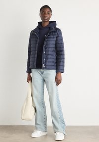 Marineblå pufferjakke med vandrette sømme, kombineret med en marineblå hættetrøje, lyseblå vidde bukser og hvide sneakers. Holder en cremefarvet tote-taske.