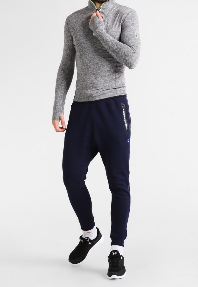 Homme portant un haut gris à manches longues zippé, un pantalon de jogging bleu marine avec poche zippée, des chaussettes blanches et des chaussures de sport noires.