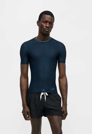 Camisa ajustada de manga corta en azul oscuro con texto "LE COL" en el cuello, combinada con pantalones cortos de baño negros con cordón blanco.