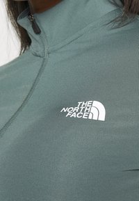 Grünes Polyester-Zip-up-Oberteil mit hohem Kragen und gerippter Textur, ausgestattet mit einem weißen Logo von "The North Face" auf der linken Brust.