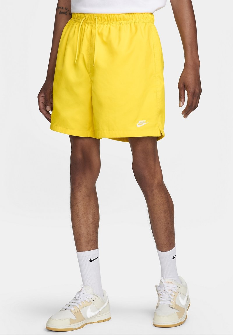 Shorts Nike jaunes vifs avec une taille élastique, un cordon de serrage et un petit logo blanc sur le devant, associés à des chaussures blanches et crème.
