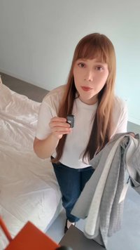 Γυναίκα με μακριά μαλλιά κρατά τηλεχειριστήριο DJI και φορά ρούχα ανοιχτού χρώματος, στέκεται κοντά σε ένα κρεβάτι σε έναν λιτά επιπλωμένο χώρο.