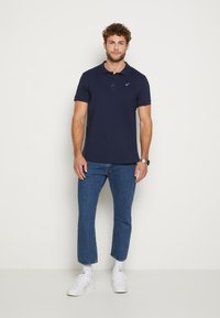 Kaiserblaue Poloshirt mit Kragen und kurzen Ärmeln, ausgestattet mit einem dezenten Logo. Kombiniert mit hellblauen Jeans und weißen Sneakers.