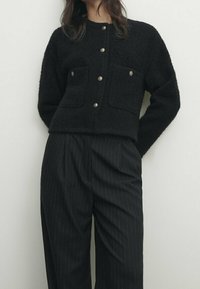 Svart, strukturerad tröja med en kortad design, två bröstfickor och rund halsringning, parat med högtalande byxor i randigt mönster.