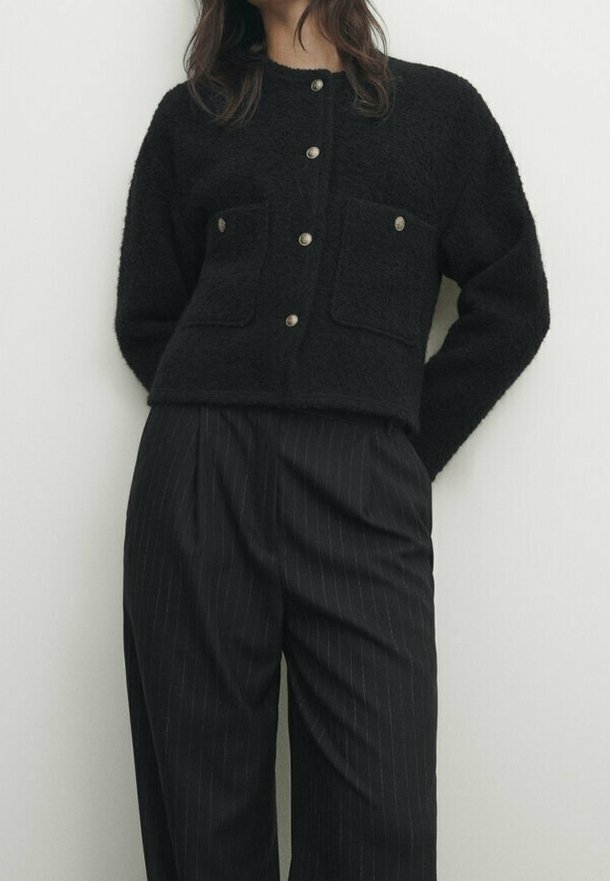 Svart, strukturerad tröja med en kortad design, två bröstfickor och rund halsringning, parat med högtalande byxor i randigt mönster.