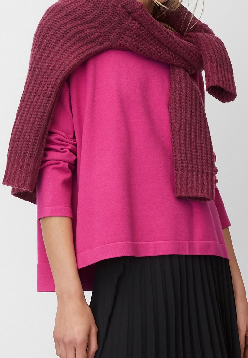 Personne portant un haut à manches longues rose vif avec un pull en maille bordeaux posé sur les épaules et une jupe plissée noire.