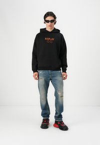 Czarna bluza z kapturem z pomarańczowym logo "REPLAY", podarte niebieskie dżinsy i czarno-czerwone masywne sneakersy. Model nosi czarne okulary przeciwsłoneczne.