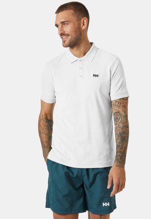 Camisa polo blanca con un acabado texturizado, tres botones y un logo negro en el lado izquierdo del pecho; combinada con pantalones cortos en color verde azulado.