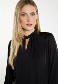 Schwarze Bluse mit hohem Kragen und Faltendetail, mit einer Schlüsselöffnung am Kragen. Glattes, leichtes Material.