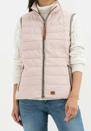 Kvinde iført en lyserød quiltet vest uden ærmer over en hvid kabelstrikket sweater og blå jeans, stående mod en hvid baggrund.