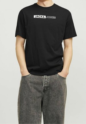 Osoba ubrana w czarną koszulkę z napisem „JACK & JONES” i wyblakłe czarne dżinsy, ręce w kieszeniach, jednolite jasne tło.
