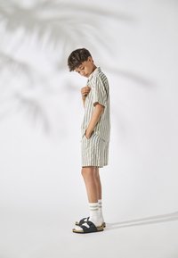 Boy wearing green-and-white striped short-sleeve shirt and shorts, standing with one hand in pocket and one hand near chest, looking down.