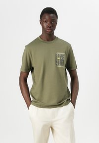T-shirt en coton vert olive à manches courtes, décolleté rond et poche poitrine avec un design imprimé de lignes et de détails textuels.