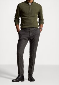 Olivgrüner Zip-Up-Pullover aus Strickstoff, kombiniert mit grauen strukturierten Hosen. Schwarze Schnürstiefel runden das Outfit mit einem eleganten Design ab.