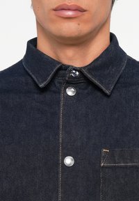 Temno denim srajca z usnjatim ovratnikom, vidnimi šivi, osrednjim zapiranjem na gumb in žepom na prsih s kontrastno nitjo.