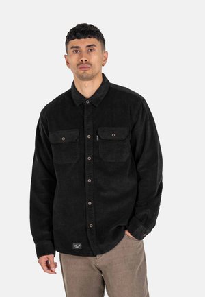 Black corduroy button-up shirt with two front pockets and copper snap buttons, worn with light brown pants, featuring a small logo label.