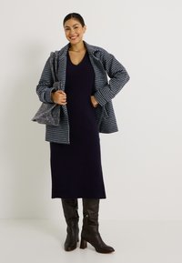 Robe tricotée côtelée marine, manteau à motif pied-de-poule, sac à main gris et bottes marron jusqu'au genou avec des talons carrés complètent la tenue.
