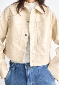 Kobieta ubrana w krótką beżową skórzaną kurtkę z zatrzaskami oraz niebieskie dżinsy o wysokim stanie, dłonie w kieszeniach.