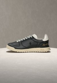 Μαύρο δερμάτινο αθλητικό παπούτσι με λευκά κορδόνια και φτέρνα, μπεζ σόλα, λογότυπο Polo στη πλευρά και υπογραφή "Ralph Lauren" σε χρυσό κοντά στη φτέρνα.