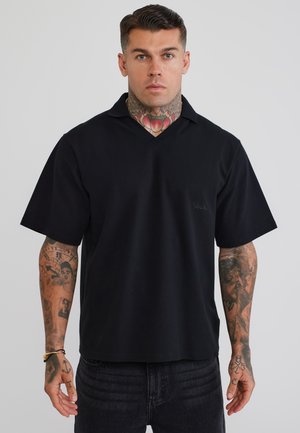 Man met tatoeages op nek en armen, gekleed in een zwart shirt met korte mouwen en kraag en donkere spijkerbroek, staand tegen een effen lichte achtergrond.