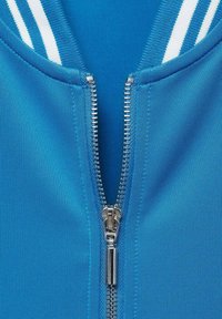 Giacca blu con zip, realizzata in tessuto liscio. Presenta una zip argentata e un colletto con strisce bianche. Inquadratura ravvicinata dell'area della zip.