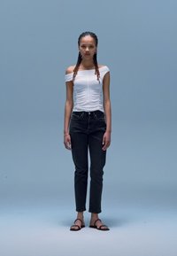 Hvit off-shoulder topp med teksturert stoff, svarte høytlivs jeans og svarte flate sandaler, satt mot en lys blå bakgrunn.