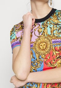 T-shirt a maniche corte con stampa multicolore vivace che presenta motivi dorati ornati e un motivo centrale a sole; bordo del colletto in nero.